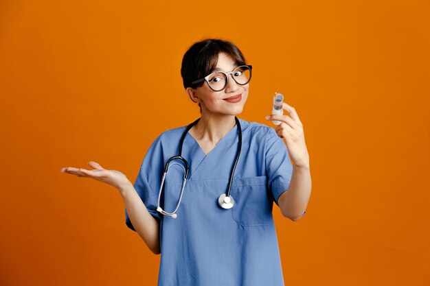 Pleased spreading hand holding syringe young female doctor wearing uniform fith stethoscope isolated on orange background