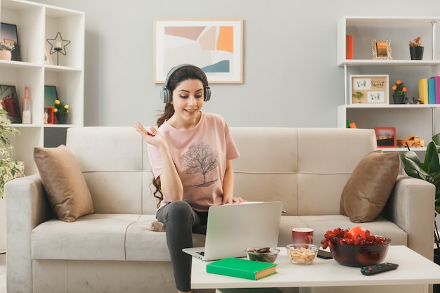 Pleased spreading hand young girl wearing headphones used laptop sitting on sofa behind coffee table in living room