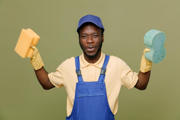 Довольный раскинув руки, держа чистящие губки, молодой афроамериканец-уборщик мужчина в униформе с перчатками, изолированными на зеленом фоне