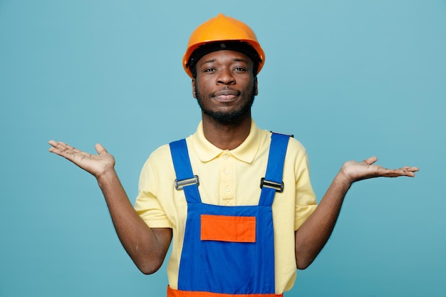 Pleased spreading hands young african american builder in uniform isolated on blue background