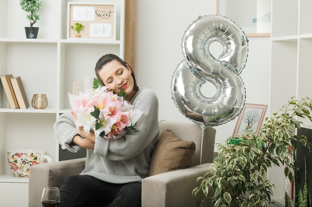 Pleased with closed eyes beautiful woman on happy women day holding bouquet sitting on armchair in living room