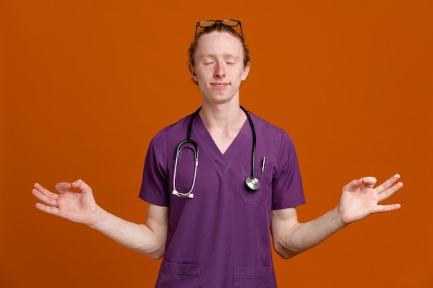 pleased with closed eyes showing meditation gesture young male doctor wearing uniform with stethoscope isolated on orange background