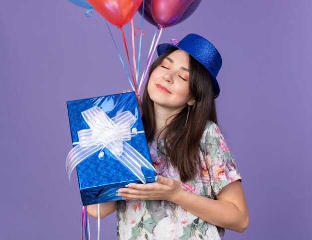 Pleased with closed eyes young beautiful girl wearing party hat holding balloons with gift box isolated on blue wall