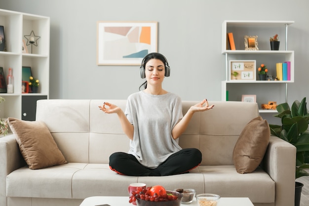 Pleased with closed eyes young girl wearing headphones and meditating on sofa behind coffee table in living room