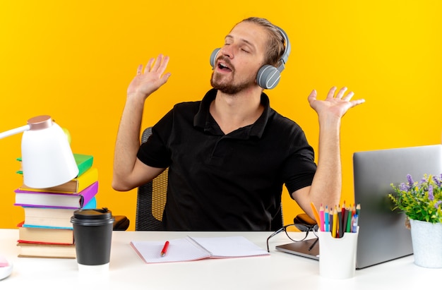 Pleased with closed eyes young guy student wearing headphones sitting at table with school tools spreading hands 