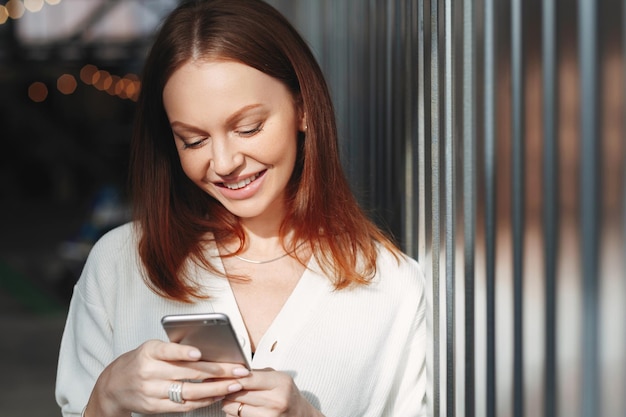 Photo pleased woman focused into screen of cell phone checks email box dressed in white clothes sends feedback connected to wireless internet has brown hair charming smile sends text message