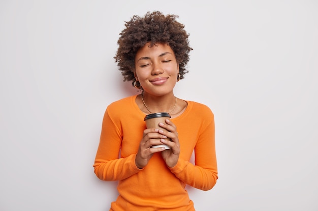 Pleased woman with curly hair holds paper disposable cup of coffee closes eyes from satisfaction wears casual orange jumper isolated over white wall