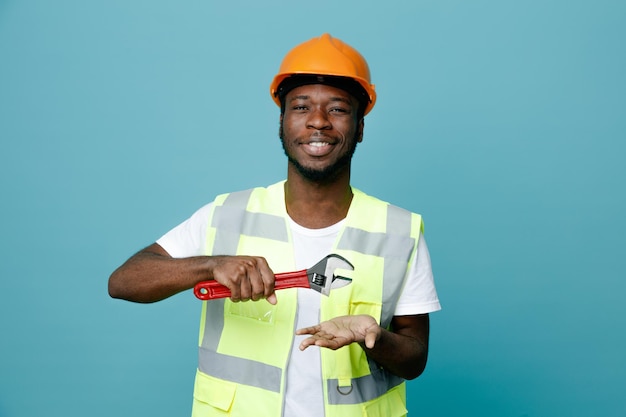 pleased young african american builder in uniform holding gas wrench isolated on blue background