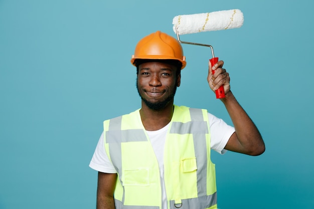 Soddisfatto giovane costruttore afroamericano in uniforme che tiene una spazzola a rullo isolata su sfondo blu