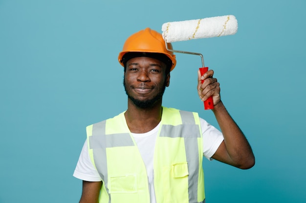 Pleased young african american builder in uniform holding roller brush isolated on blue background