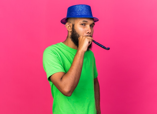 Pleased young afro-american guy wearing party hat blowing party whistle isolated on pink wall