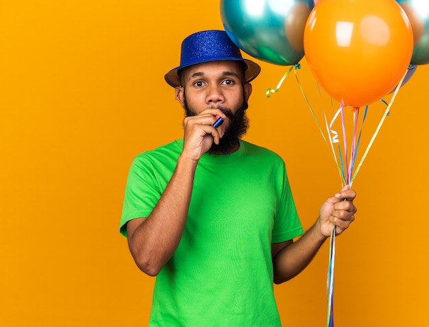 Felice giovane ragazzo afro-americano che indossa un cappello da festa con in mano palloncini che soffiano un fischio di festa