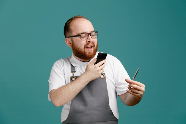 Pleased young barber wearing uniform and glasses holding scissors taking photo of it isolated on blue background