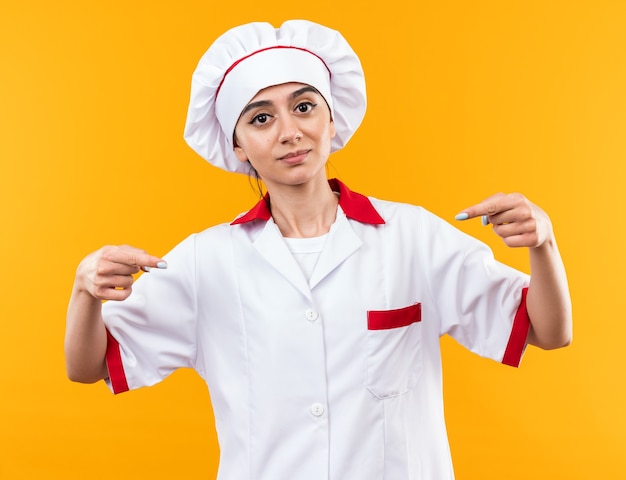 La giovane e bella ragazza soddisfatta in uniforme da chef punta su se stessa isolata sul muro arancione