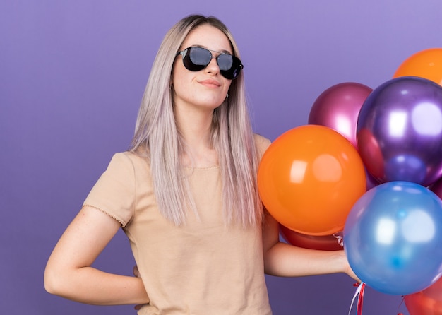 Pleased young beautiful girl wearing glasses holding balloons putting hand on hip 