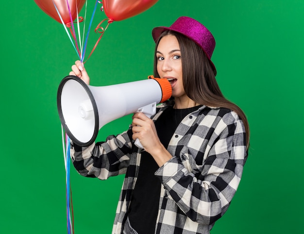 Pleased young beautiful girl wearing party hat holding balloons speaks on loudspeaker 