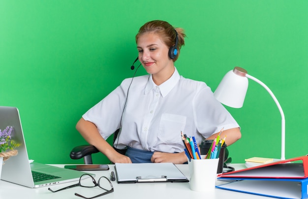 Pleased young blonde call centre girl wearing headset sitting at desk with work tools looking at laptop 