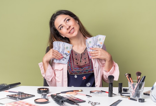 Pleased young brunette girl sitting at table with makeup tools holding money and looking up isolated on olive green wall with copy space