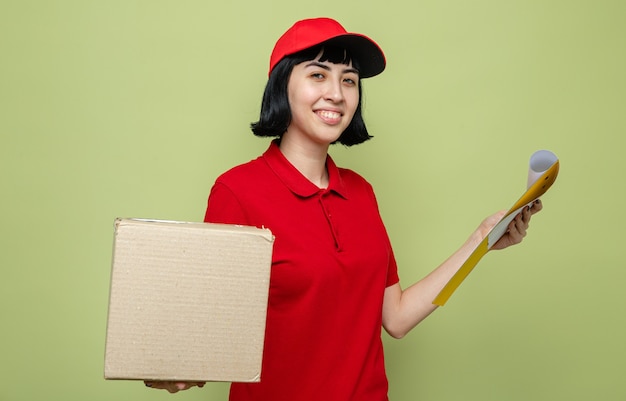 Pleased young caucasian delivery girl holding cardboard box and clipboard 