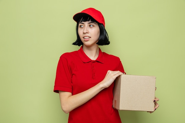 Pleased young caucasian delivery woman holding cardboard box and looking at side 