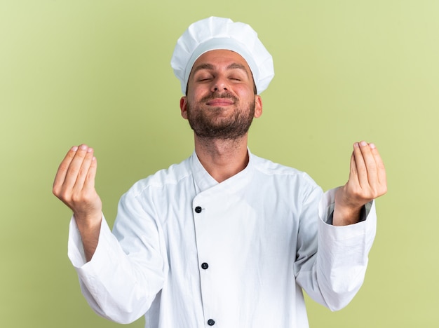 Pleased young caucasian male cook in chef uniform and cap keeping hands in yoga gesture with closed eyes isolated on olive green wall