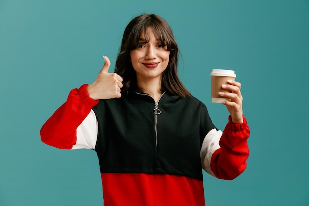 Довольная молодая кавказская женщина показывает бумажную кофейную чашку, глядя в камеру, показывая большой палец вверх на синем фоне
