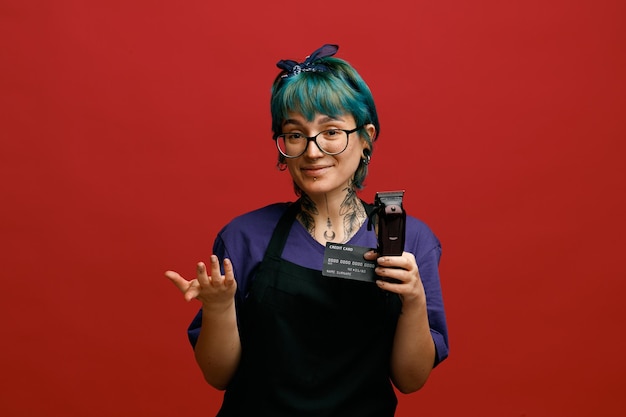 Pleased young female barber wearing uniform glasses and headband looking at camera showing credit card and hair trimmer and empty hand isolated on red background