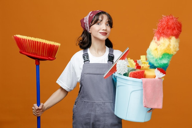 Pleased young female cleaner wearing uniform and bandana holding squeegee mop and bucket of cleaning tools looking at camera isolated on orange background