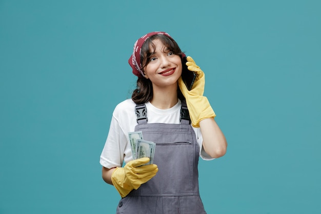 Felice giovane donna addetta alle pulizie che indossa bandana uniforme e guanti di gomma che tengono soldi guardando di lato mentre si parla al telefono isolato su sfondo blu