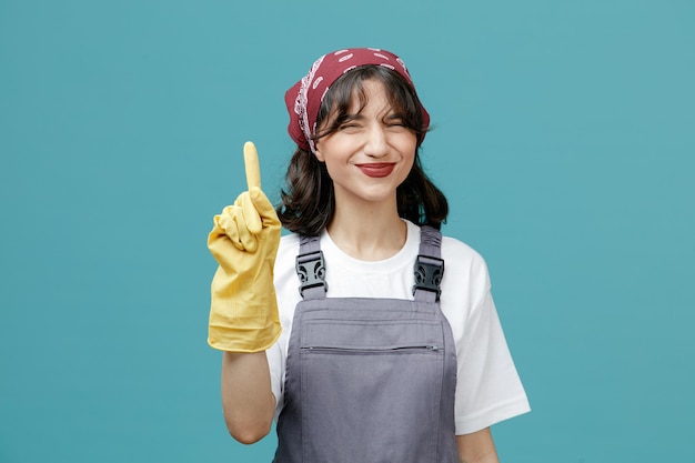 Pleased young female cleaner wearing uniform bandana and rubber gloves looking at camera pointing up isolated on blue background