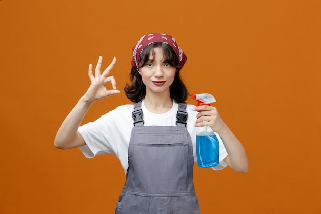 Pleased young female cleaner wearing uniform and bandana showing cleanser looking at camera showing ok sign isolated on orange background