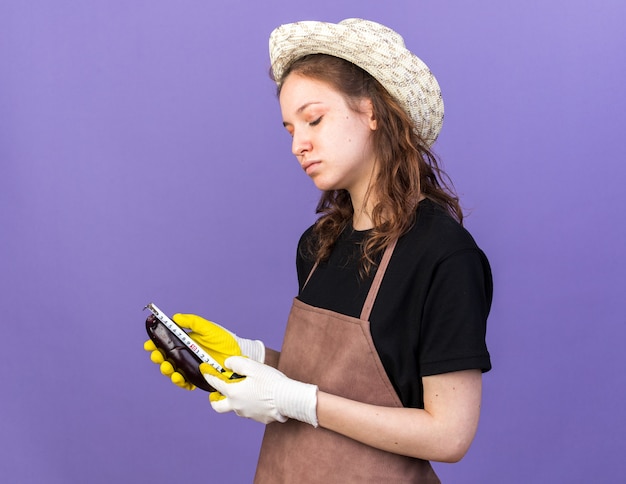 Pleased young female gardener wearing gardening hat with gloves measuring eggplant with tape measure isolated on blue wall