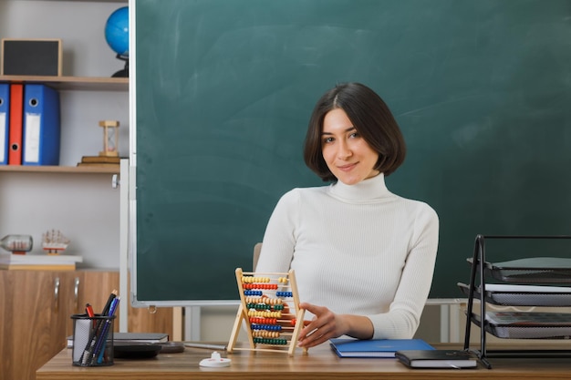 pleased young female teacher holding abacus sitting at desk with school tools on in classroom