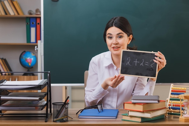 pleased young female teacher holding and points at mini blackboard sitting at desk with school tools in classroom
