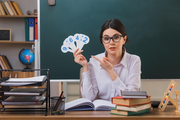 pleased young female teacher wearing glasses holding and points at number fun sitting at desk with school tools in classroom