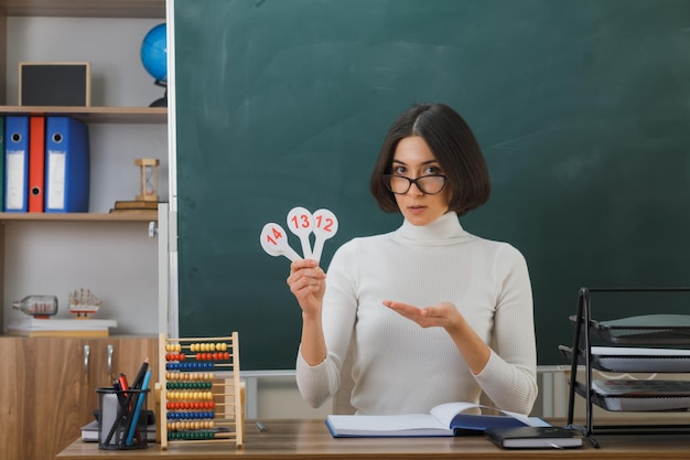 pleased young female teacher wearing glasses holding and points with hand number fan sitting at desk with school tools on in classroom