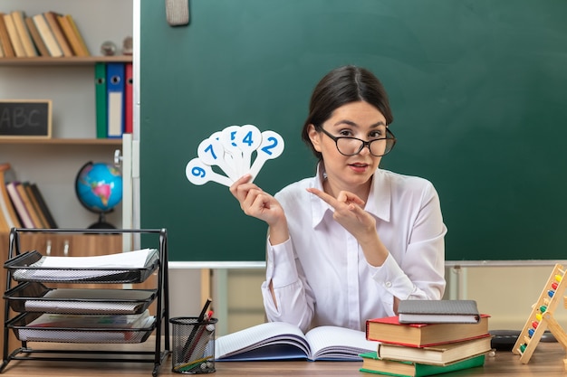 Pleased young female teacher wearing glasses sitting at table with school tools holding and points at number fans in classroom