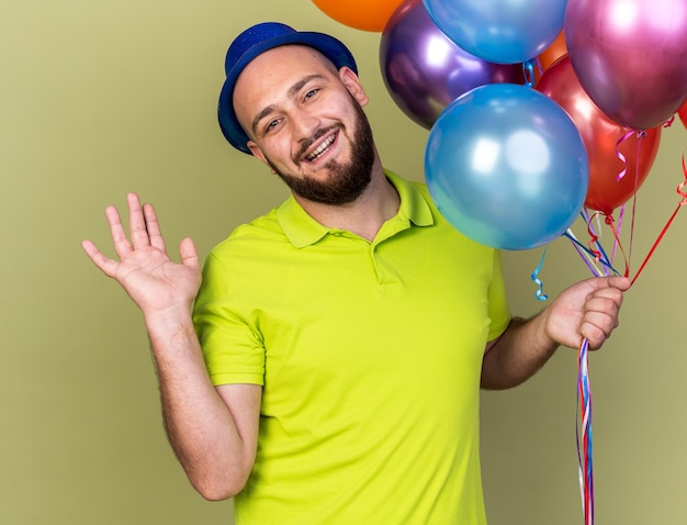 Pleased young guy wearing party hat holding balloons spreading hands isolated on olive green wall