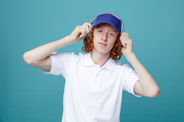 Pleased young handsome guy in cap wearing headphones isolated on blue background