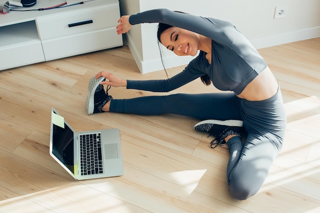 Piacevole giovane donna in abiti sportivi che allunga le gambe davanti a un laptop e sorride mentre si piega verso il piede