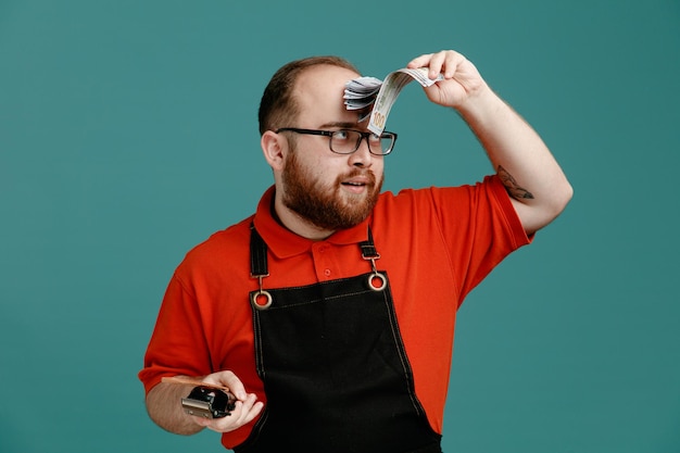 Pleased young male barber wearing glasses red shirt and barber apron holding comb hair clipper touching forehead with money looking at money isolated on blue background