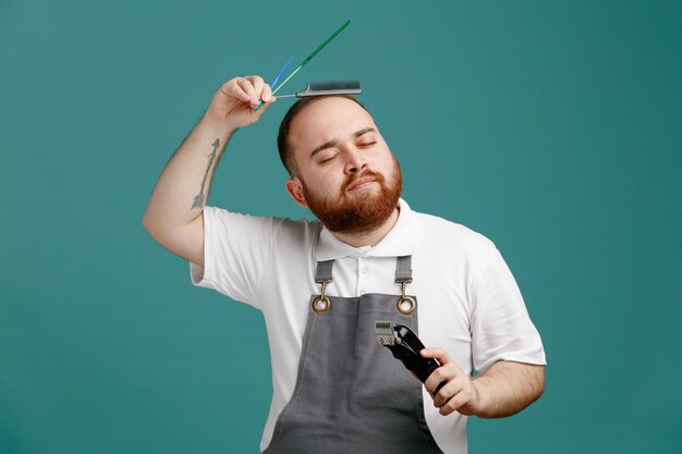 Pleased young male barber wearing white shirt and barber apron holding hair trimmer and combs touching head with comb with closed eyes isolated on blue background