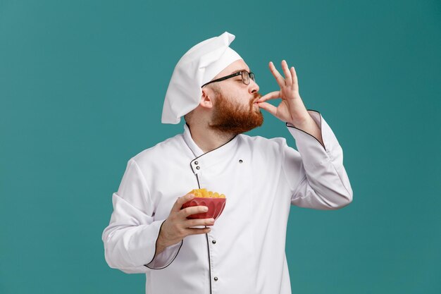 Pleased young male chef wearing glasses uniform and cap holding bowl of macaronis turning head to side making delicious gesture with closed eyes isolated on blue background