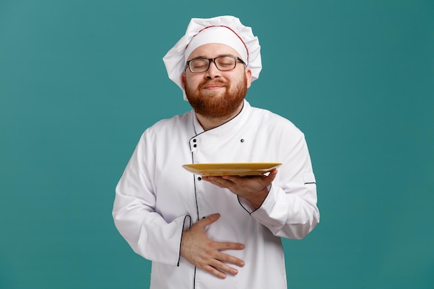 Pleased young male chef wearing glasses uniform and cap holding empty plate showing hungry gesture with closed eyes isolated on blue background