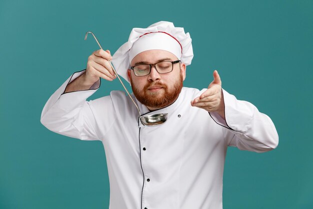 Pleased young male chef wearing glasses uniform and cap holding ladle keeping hand in air pretend smelling aroma of meal with closed eyes isolated on blue background