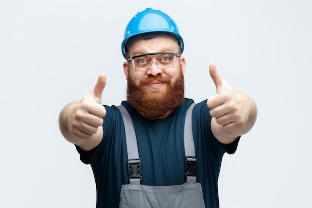 Pleased young male construction worker wearing safety helmet uniform and safety glasses looking at camera stretching hands out towards camera showing thumbs up isolated on white background