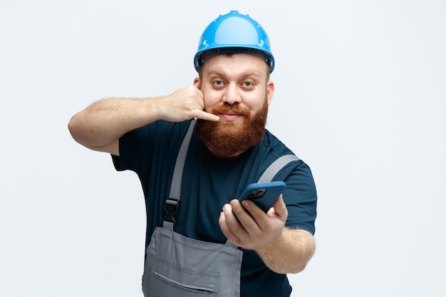 Felice giovane operaio edile maschio che indossa un casco di sicurezza e uniforme che allunga il telefono cellulare verso la fotocamera che mostra il gesto di chiamata isolato su sfondo bianco