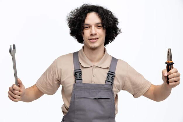 Pleased young male construction worker wearing uniform holding spanner and pliers while looking at camera isolated on white background