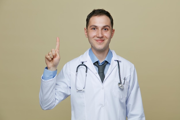 Pleased young male doctor wearing medical robe and stethoscope around neck looking at camera pointing up isolated on olive green background