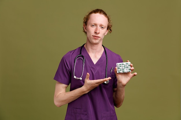 Pleased young male doctor wearing uniform with stethoscope holding and points at pills isolated on green background